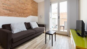 Ein modernes Wohnzimmer mit einer Ziegelwand, einer Couch mit gemusterten Kissen, einem kleinen Tisch, einem Fernseher auf einem grünen Schrank und einer Glastür, die zu einem Außenbereich führt.