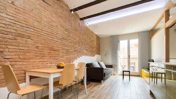 Ein stilvoller Wohnraum mit Backsteinwand, einem Essbereich mit Tisch und Stühlen, einer Couch und einem Balkon mit Geländer.