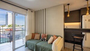 Un apartamento moderno con balcón, un sofá con cojines, una zona de cocina con una lámpara colgante y vistas a un paisaje urbano.
