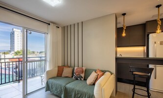 Un apartamento moderno con balcón, un sofá con cojines, una zona de cocina con una lámpara colgante y vistas a un paisaje urbano.