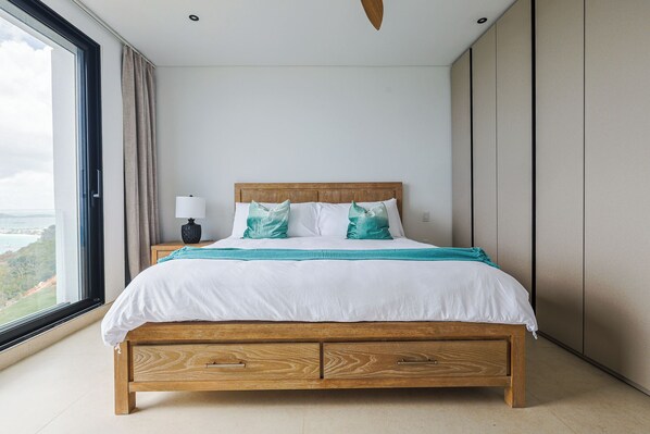 A bedroom with a wooden bed, white bedding, teal pillows, a nightstand with a lamp, and a large window with a view. - Villa Turquoise Heaven (Sint Maarten)