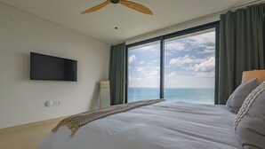 A bedroom with a large bed, a ceiling fan, a TV mounted on the wall, and a view of the ocean through a large window. - Villa Turquoise Heaven (Sint Maarten)