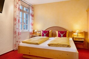 Un lit en bois avec deux oreillers, une table de chevet avec une lampe, une fenêtre avec des rideaux à motifs floraux et un tapis rouge.