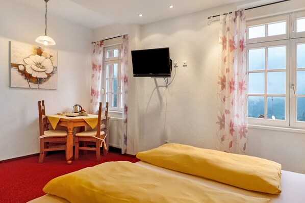 Une pièce comprenant un lit, une table avec des chaises, un téléviseur fixé au mur et une fenêtre avec des rideaux.