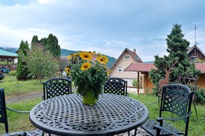 Une table ronde en métal, ornée d’un pot de fleurs au centre, entourée de chaises en métal, installée à l’extérieur, avec des maisons et des arbres en arrière-plan.