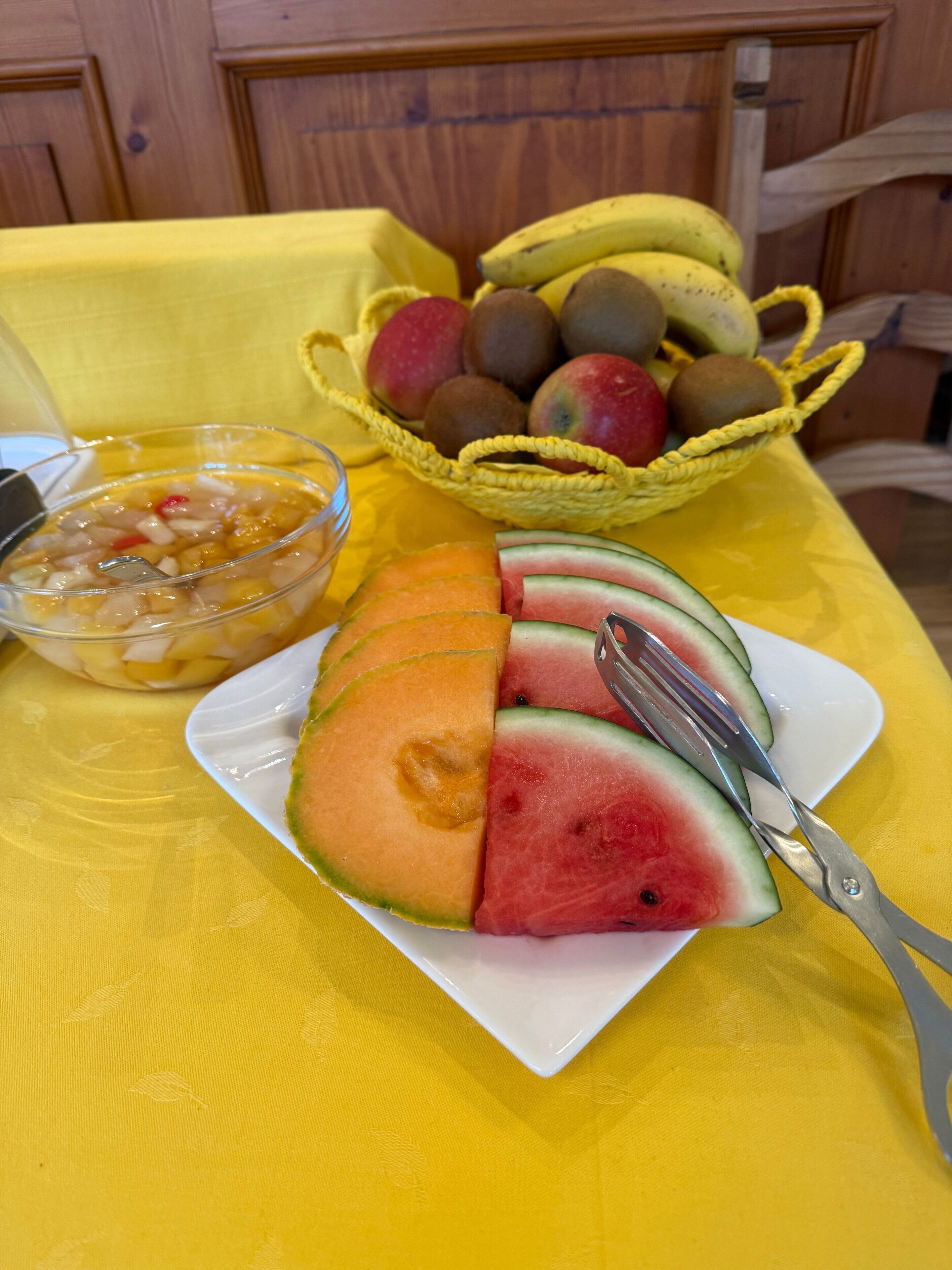 A table with a yellow tablecloth, a plate of sliced watermelon and cantaloupe, a bowl of fruit salad, and a basket of kiwis and apples.