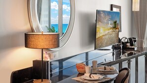 Habitación de hotel con un espejo grande, un cuadro enmarco de Miami Beach, una mesa de vidrio con alimentos para el desayuno y vista al océano a través de la ventana.