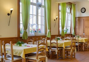 Un espace repas avec des tables et des chaises en bois, des rideaux verts et une horloge au mur.