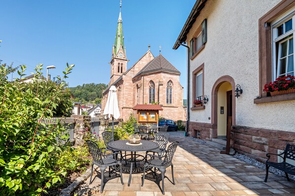 Une cour avec une table ronde et des chaises, un banc, une église dotée d’un clocher imposant et un bâtiment avec une fenêtre et une jardinière.