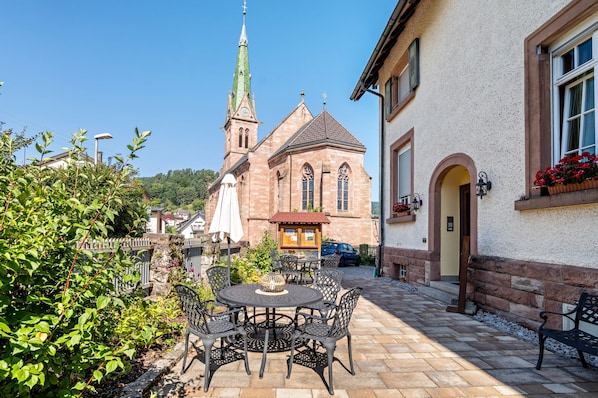 A courtyard with a round table and chairs, a bench, a church with a tall steeple, and a building with a window and flower box.
