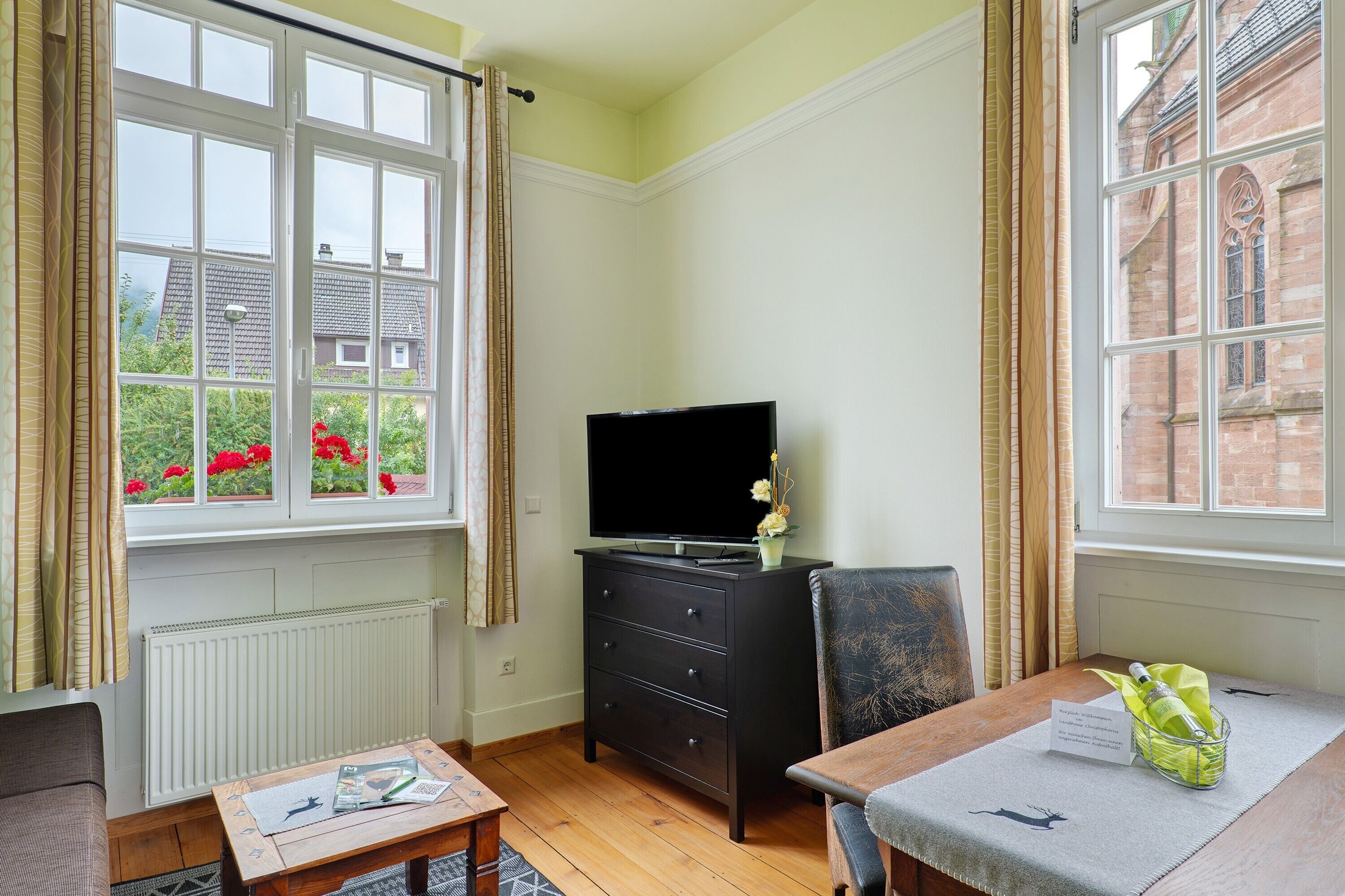 A room with a TV on a dresser, a table with a name card, and a window with curtains.