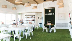 A dining area with white tables and chairs, a sign reading "PICNIC BEACH", and a bookshelf in the background. - Beautiful 2B/2B Beach Views & Best Eats | Palmaway (Gulf Shores)