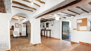 A rustic interior with exposed wooden beams, white walls, and a tiled floor. There are two white chairs and a table with a plant.