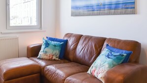 A brown leather sofa with two decorative pillows, a window with a view of buildings, and a painting of a beach scene on the wall. - 3 bedroom accommodation in Ilfracombe (Ilfracombe)