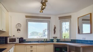 A kitchen with a microwave, sink, and bar stool. There are windows with views of buildings and a clock on the wall. - 3 bedroom accommodation in Ilfracombe (Ilfracombe)