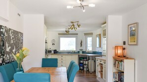 A modern kitchen with white cabinets, a wooden dining table, teal chairs, and a central island. - 3 bedroom accommodation in Ilfracombe (Ilfracombe)