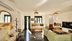 A spacious living area with a sofa, a dining table, and a TV. There is a mirror on the wall and a door leading to another room.