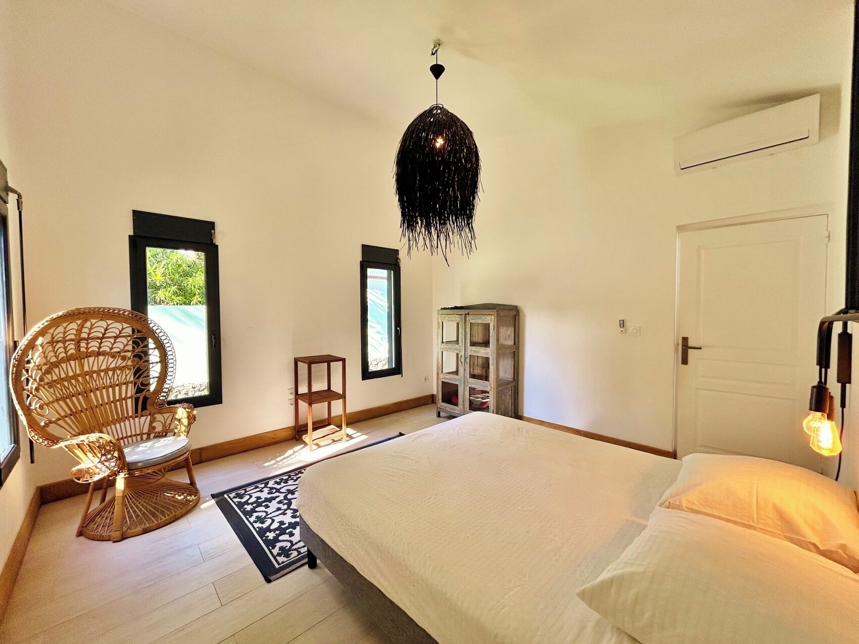 Une chambre à coucher avec un grand lit, une chaise en osier, une petite table, une armoire et un luminaire suspendu.