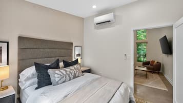 A modern bedroom with a large bed, a nightstand, an air conditioning unit, a television, and a view of a green outdoor area.