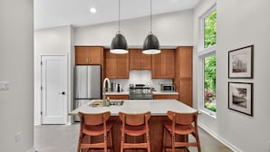 A modern kitchen with wooden cabinets, a large island with bar stools, stainless steel appliances, pendant lighting, and framed pictures on the wall. - 500 House - Unit A 1 Bedroom Townhouse (Mequon)