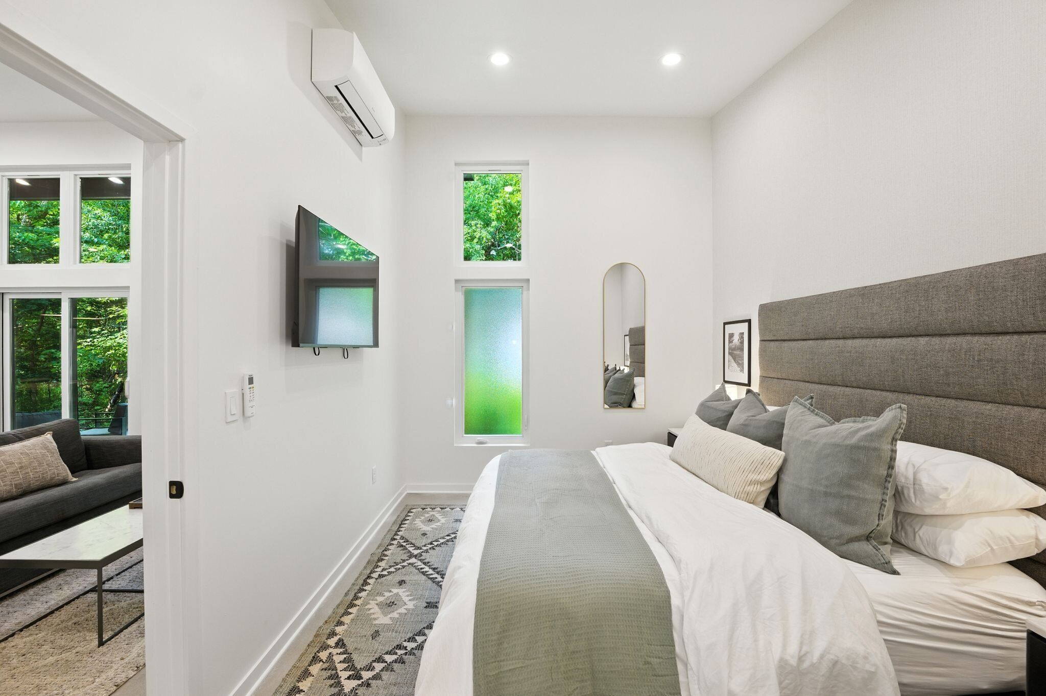 A modern bedroom with a large bed, a grey headboard, a TV mounted on the wall, and a window with a view of greenery.