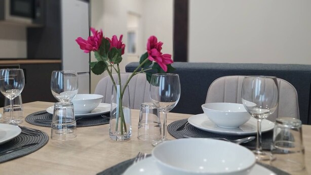 Une table à manger dressée avec de l’assiette blanche, des bols et des verres à vin, agrémentée de deux vases remplis de fleurs roses.