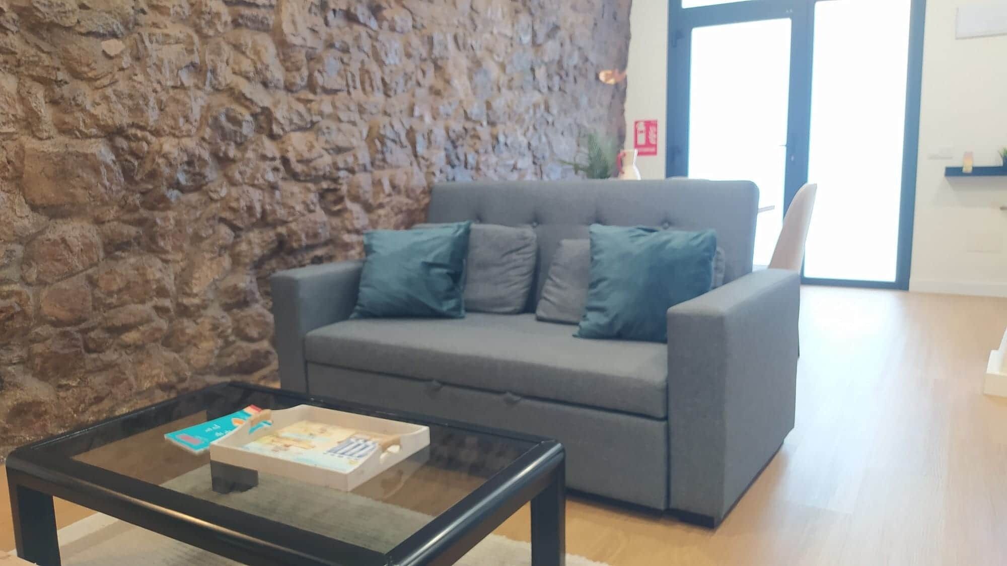 A modern living room with a stone wall, grey sofa, and a glass coffee table with a book.