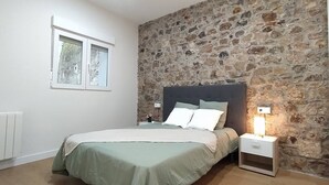 Une chambre avec un mur en pierre, un lit recouvert d’une couverture verte, deux tables de chevet avec des lampes et une fenêtre donnant sur des arbres.