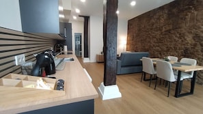 A modern kitchen with a kettle, a coffee machine, and a wooden countertop. There is a dining area with a table and chairs, and a sofa.