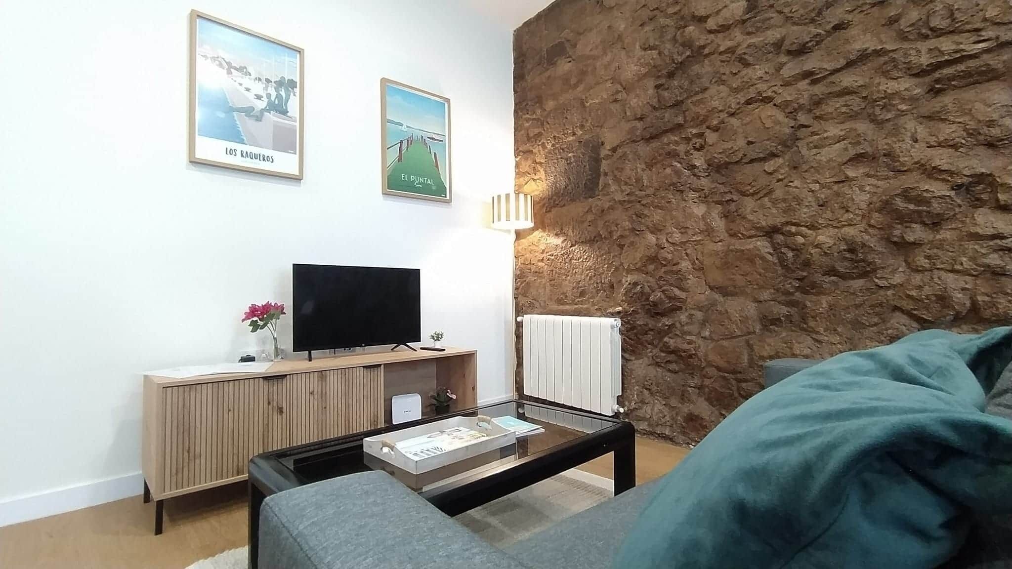 En stue med en stenvæg, et fladskærms-tv på et træbord, en sofa og et sofabord med en bog.