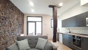 Un salon moderne avec un mur en pierre, un canapé gris et une cuisine dotée de meubles sombres et d’un four encastré.
