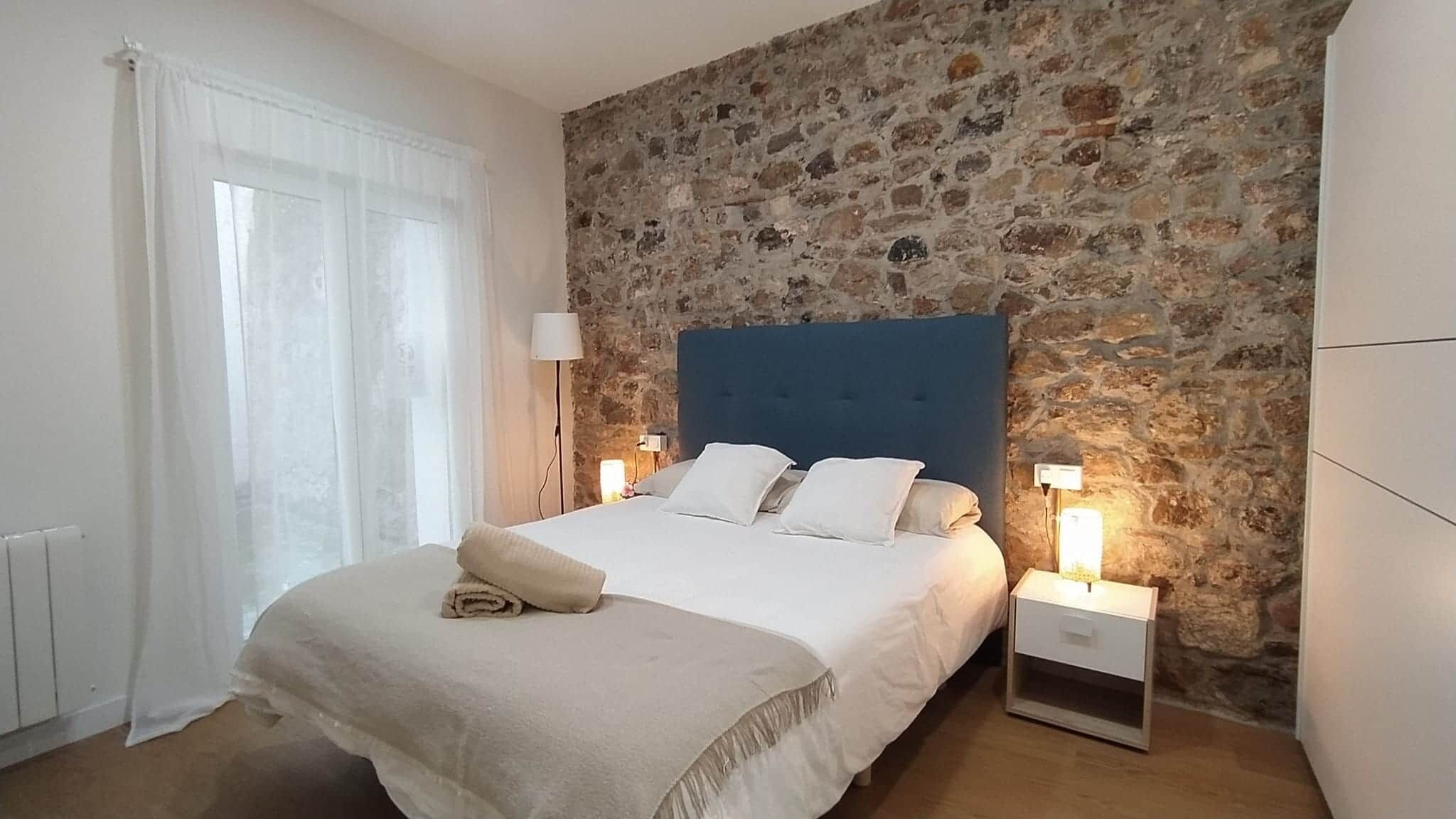 A bedroom with a stone wall, a bed with white bedding, two bedside tables with lamps, and a window with sheer curtains.