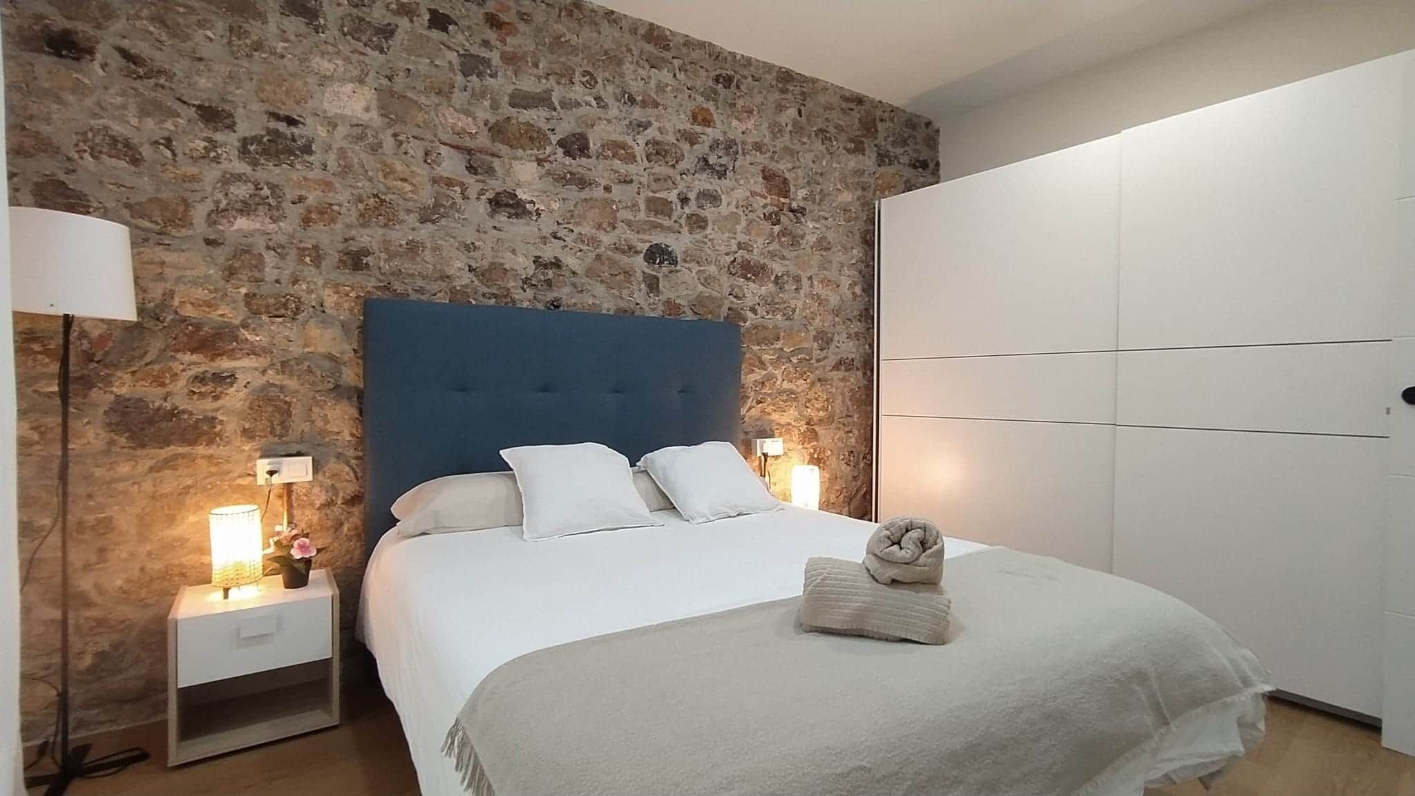 A bedroom with a stone wall, a blue headboard, a bed with white bedding, a nightstand with a lamp, and a wardrobe.