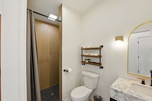 A bathroom with a toilet, a sink with a marble countertop, a shower with a curtain, and a shelf with toiletries. - 500 House - Unit C 1 Bedroom Townhouse (Mequon)