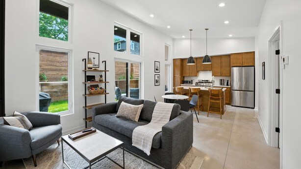 A modern living room with a grey sofa, a coffee table, and a bookshelf. There is a kitchen area with wooden cabinets and stainless steel appliances. - 500 House - Unit D 2 Bedroom Townhouse (Mequon)