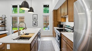 A modern kitchen with a central island, stainless steel appliances, wooden cabinets, and large windows offering views of greenery. - 500 House - Unit D 2 Bedroom Townhouse (Mequon)