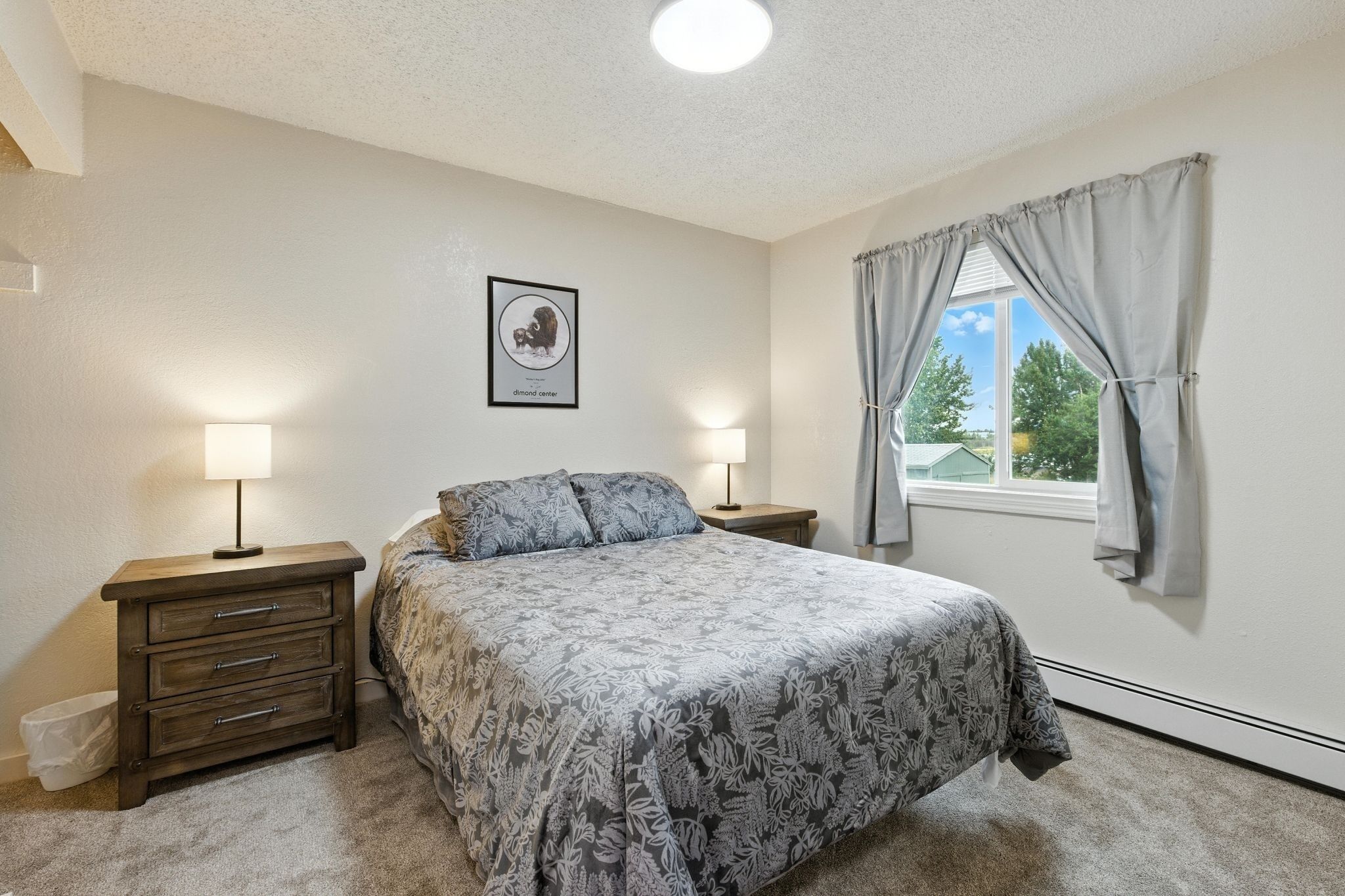 Ein Schlafzimmer mit einem Bett, zwei Nachttischen, einem Wandbild und einem Fenster mit Vorhängen.