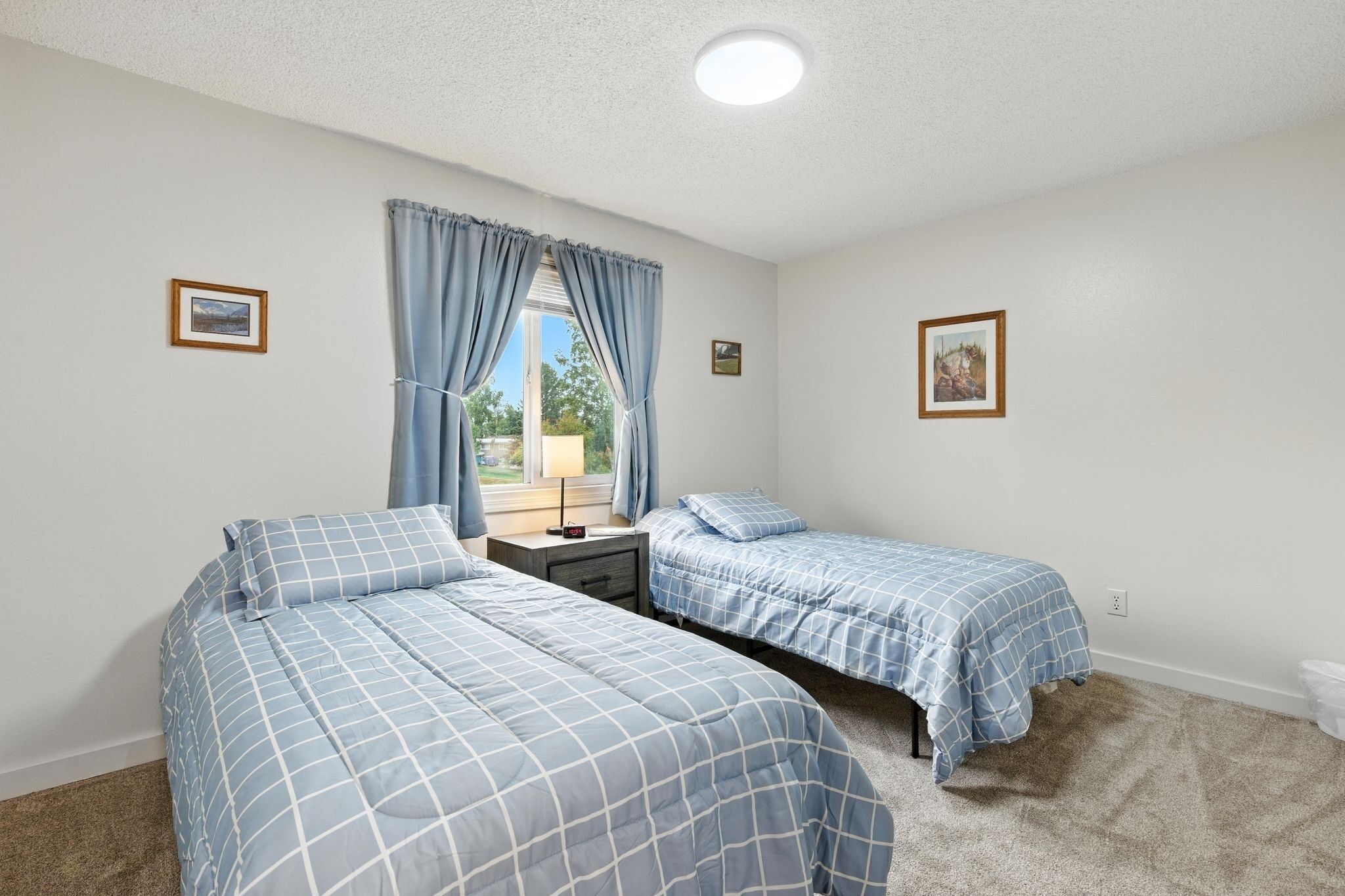 A bedroom with two beds, a window with curtains, a nightstand, and a picture on the wall.