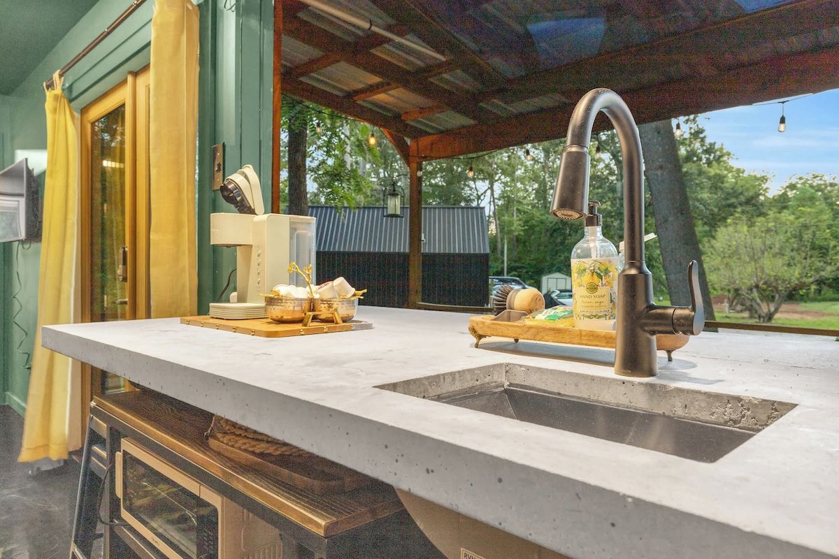 A kitchen with a large countertop, a sink, and a hanging faucet. There are cabinets below the countertop and a window with curtains.