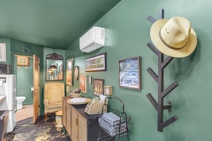 A room with a green wall, a hat rack, framed pictures, a vintage chair, a small table with a basket, and a white air conditioner. - The Sassafras - Shipping Container, shared Hot Tub (Easley)