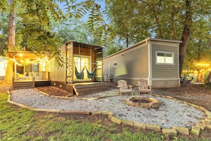 A campsite with a small wooden structure and a larger mobile home, both surrounded by trees and gravel.