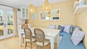 Una cocina moderna con armarios blancos, una mesa de comedor y un sofá azul. Hay lámparas colgantes de mimbre y un espejo grande sobre el fregadero.