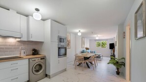 A modern kitchen with white cabinets, a built-in oven, and a washing machine. There is a dining area with a table and chairs, and a TV mounted on the wall.