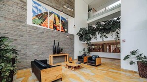 Eine großzügige Lobby mit hoher Decke, einer Steinsäule und einem großen Fenster mit Blick auf ein farbenfrohes Gebäude. Mehrere schwarze Ledersofas, ein Holztisch und ein Topf mit Pflanze sind vorhanden.