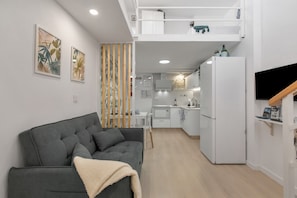 A modern living room with a grey sofa, a kitchen in the background, and a white refrigerator.