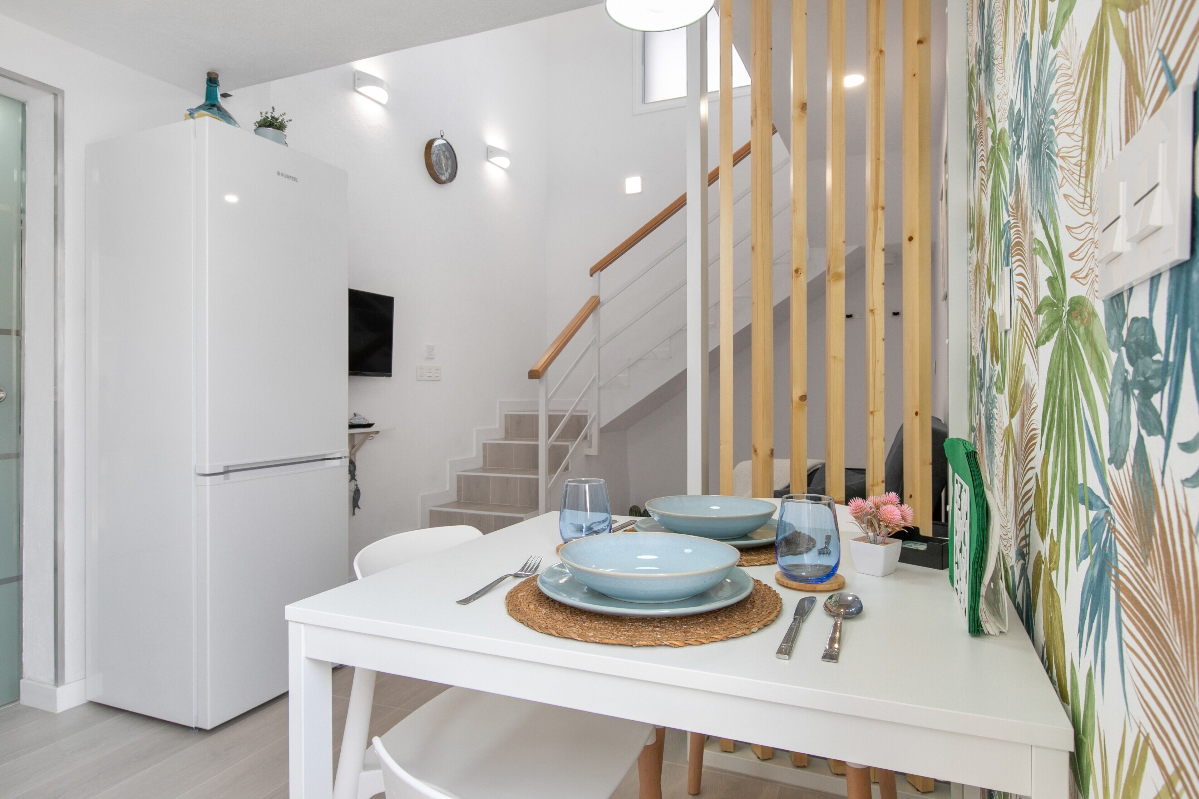 A modern kitchen with white appliances, a dining table set with blue plates, and a staircase with wooden railings.