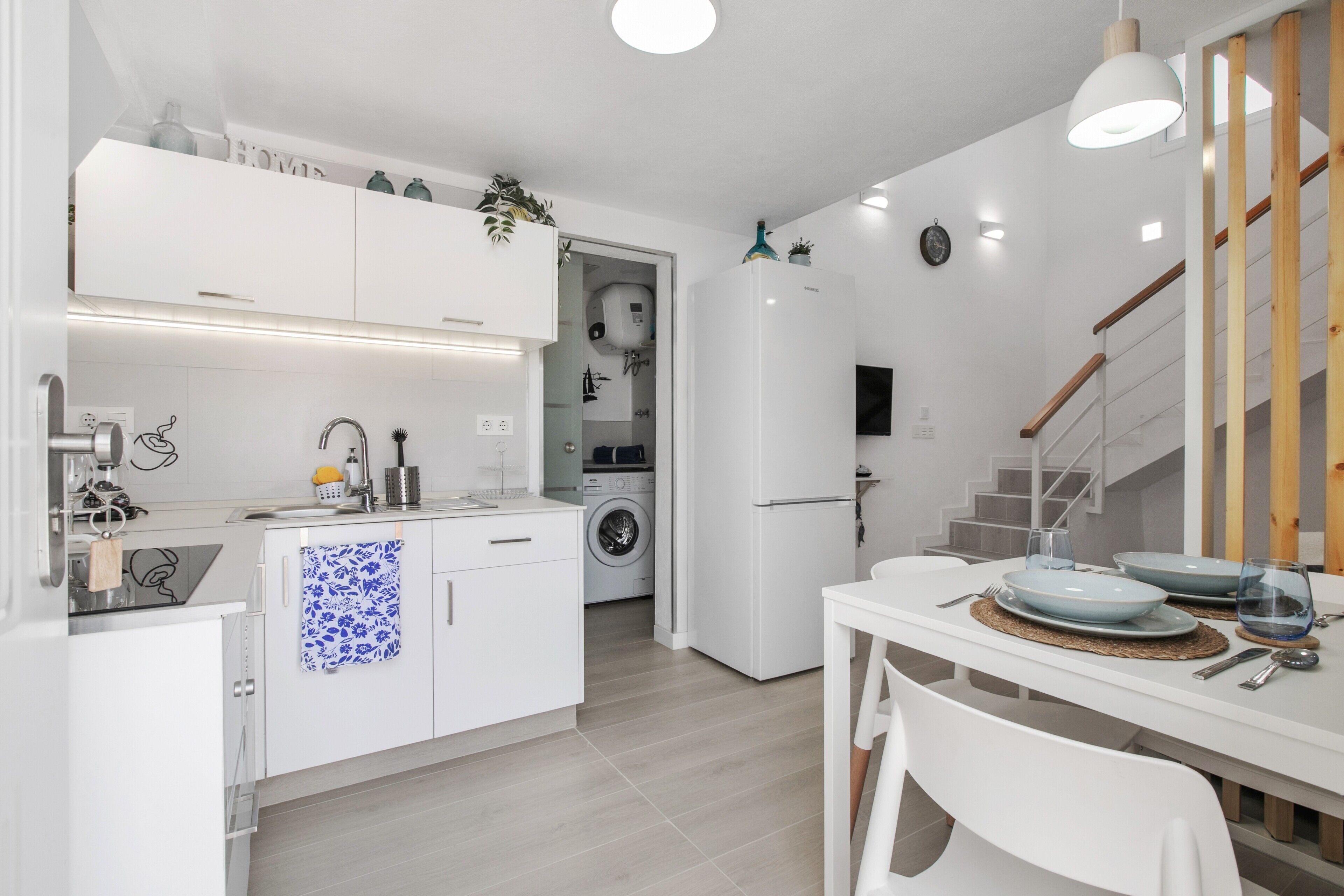 Una cocina moderna con muebles blancos, lavadora incorporada y un comedor con una mesa preparada para dos.