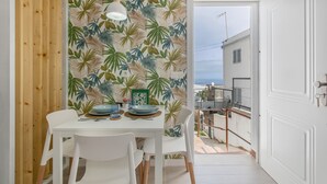 A dining area with a tropical leaf-patterned wall, white furniture, and a view of the sea through an open door.