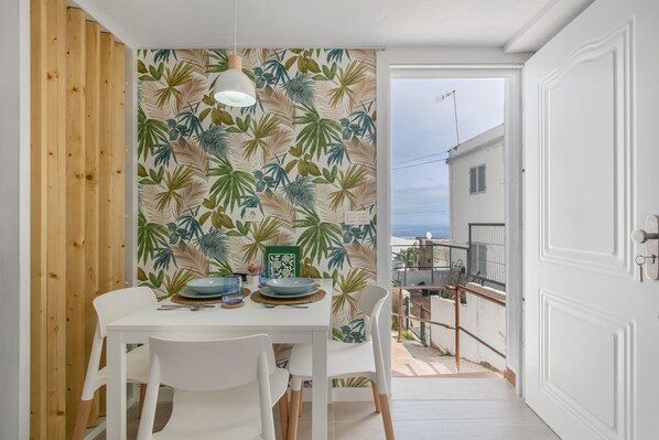 A dining area with a tropical leaf-patterned wall, white furniture, and a view of the sea through an open door.