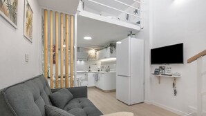 A modern living room with a grey sofa, a white TV mounted on the wall, and a kitchen in the background.